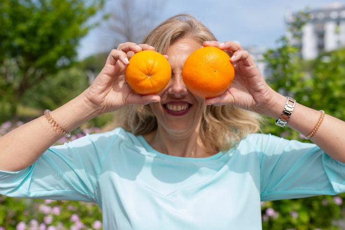 A smiling woman playfully holding oranges in a sunny park setting.