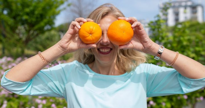A smiling woman playfully holding oranges in a sunny park setting.