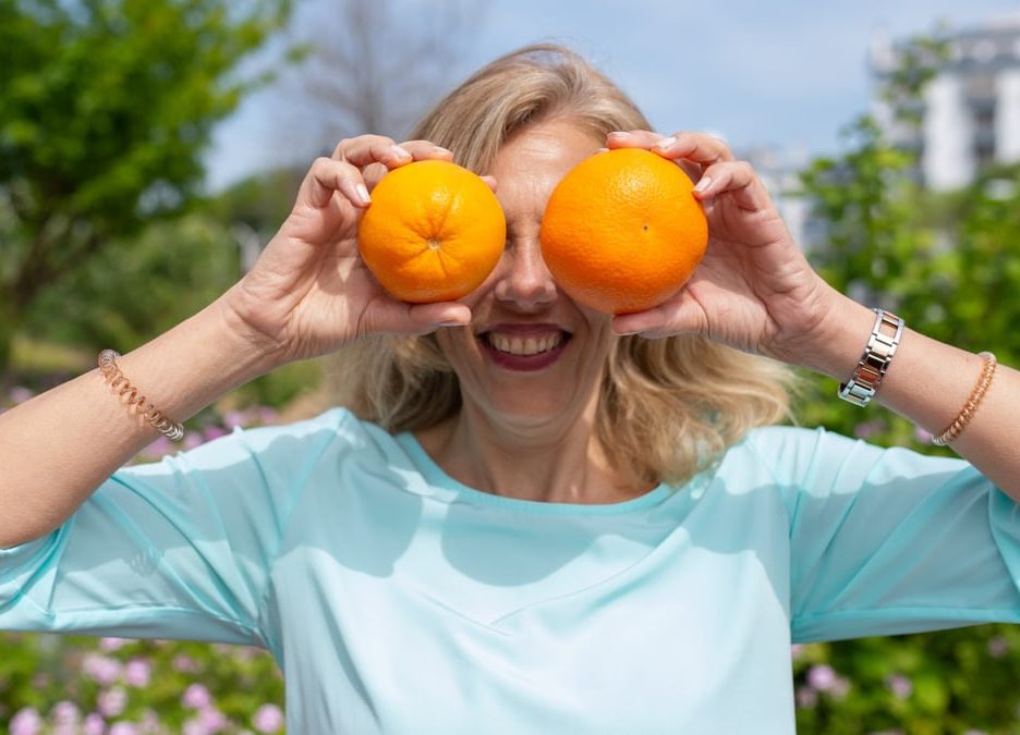 A smiling woman playfully holding oranges in a sunny park setting.